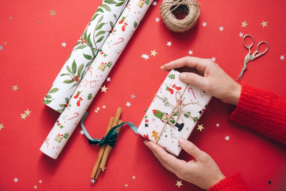 Red background with reflective stars on the table. There are 2 rolls of wrapping paper, a ball of twine and scissors sitting on the table. With hands holding a wrapped present.